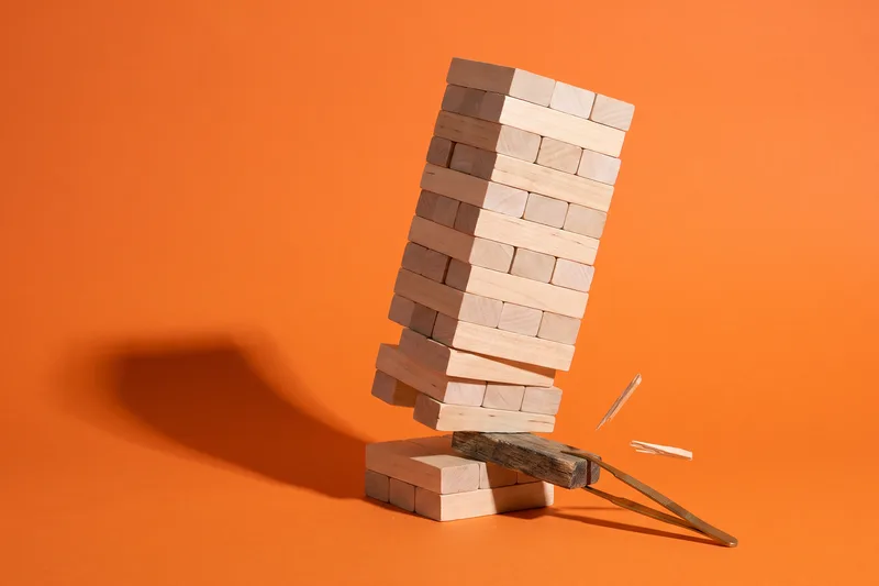Ein Turm aus hellen Holzklötzen vor orangefarbenem Studiohintergrund: Ganz unten zieht eine Messingpinzette einen alten, dunklen Block heraus, der Turm beginnt zu kippen, zwei Holzsplitter fliegen