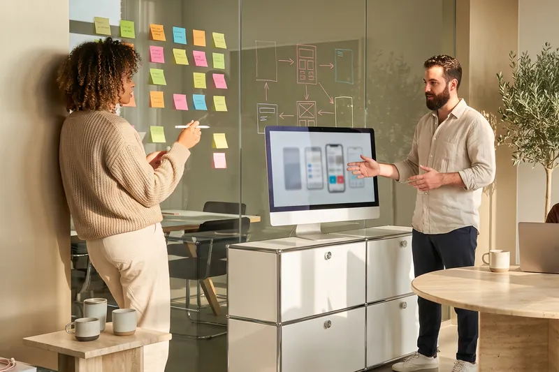 Illustration: A woman writes sticky notes on a glass wall while a man explains a workflow at a display