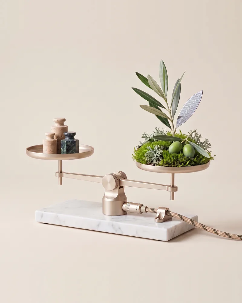 Precision balance in champagne metal on white Calacatta: weights of travertine and Verde marble on the left, a cushion of moss with lichen, an olive branch, a silicon leaf and two green olives on the right; a braided cable plugs into the marble base